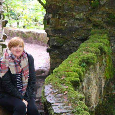 Photo of Rebecca sitting under a stone arch, near a moss-covered stone wall in Scotland, looking enchanted by her dreamy surroundings.